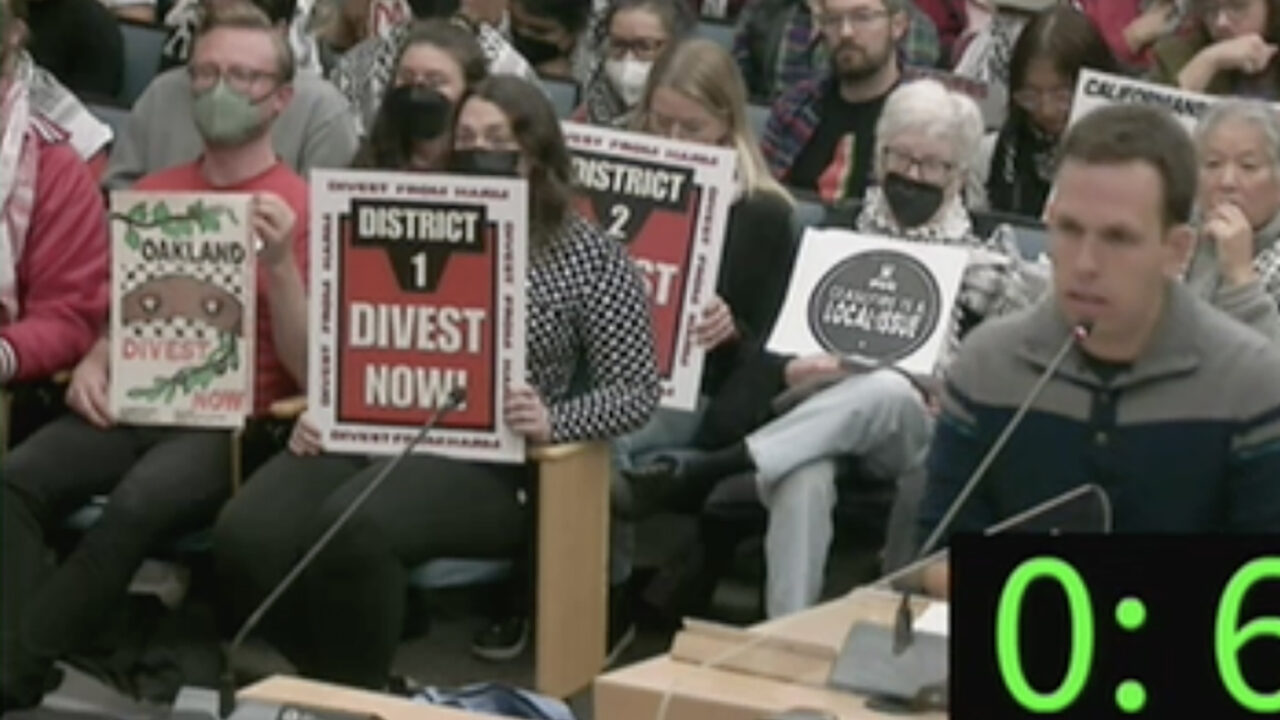 A man speaks at a lectern. Behind him, "Divest Now" signs can be seen among a seated crowd.